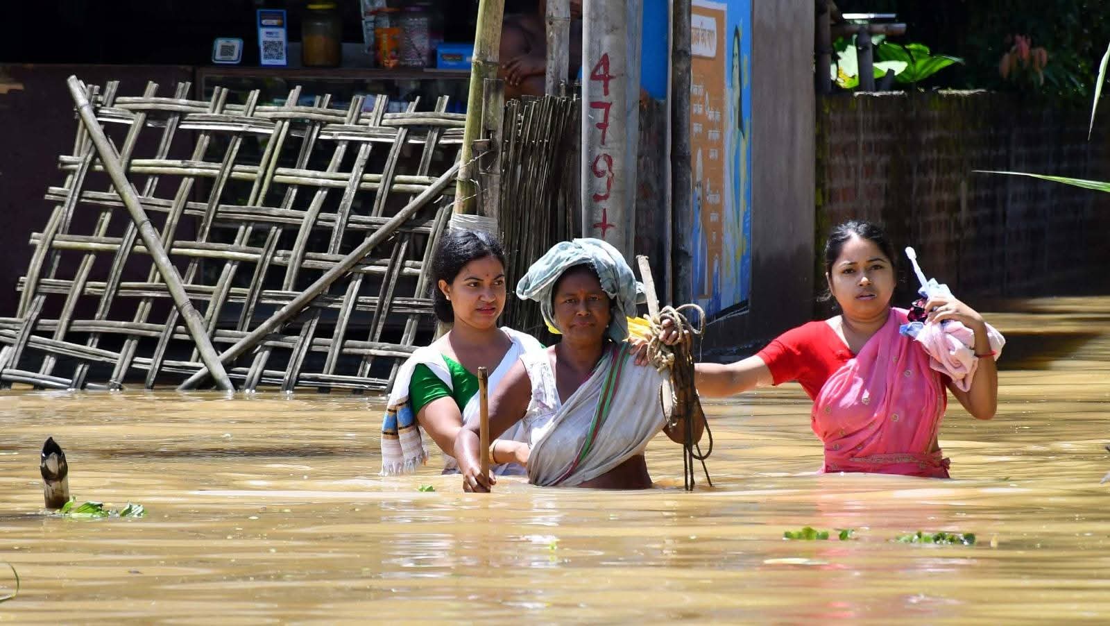 Three women wade chest-deep through muddy floodwater past a submerged village shopfront, carrying belongings above the waterline